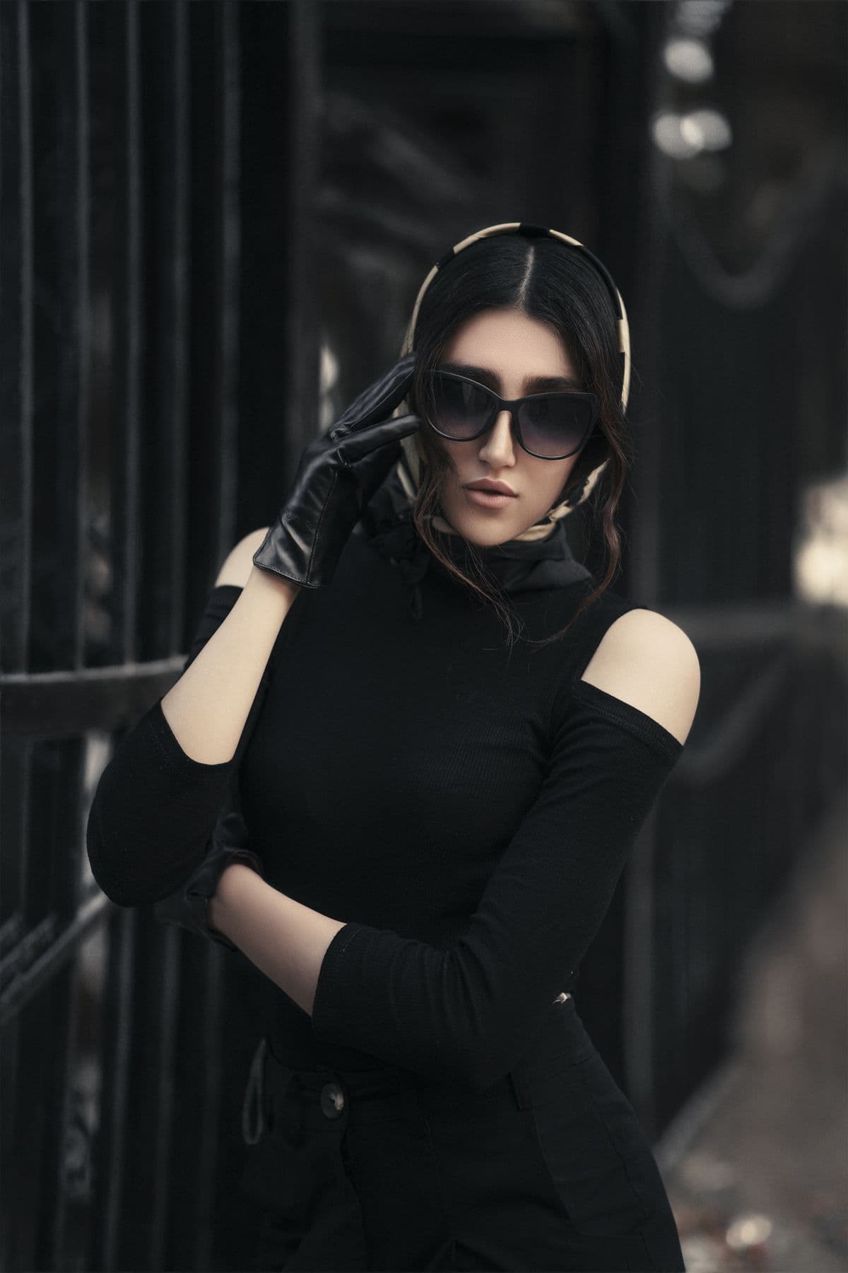 Woman in a black top, headscarf and sunglasses posing against dark railings